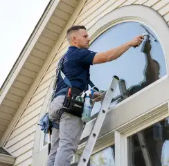 Professional window cleaner on a ladder using a squeegee to clean an exterior arched window, Grand Rapids, MI.